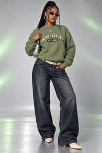 A woman modeling an olive green sweatshirt with 'WORD CARRIERS EST 2020 WILD TASTH SINCE 2020' printed on the front, paired with wide-leg dark blue jeans, beige shoes, and sunglasses, set against a gray studio background with green light flares.