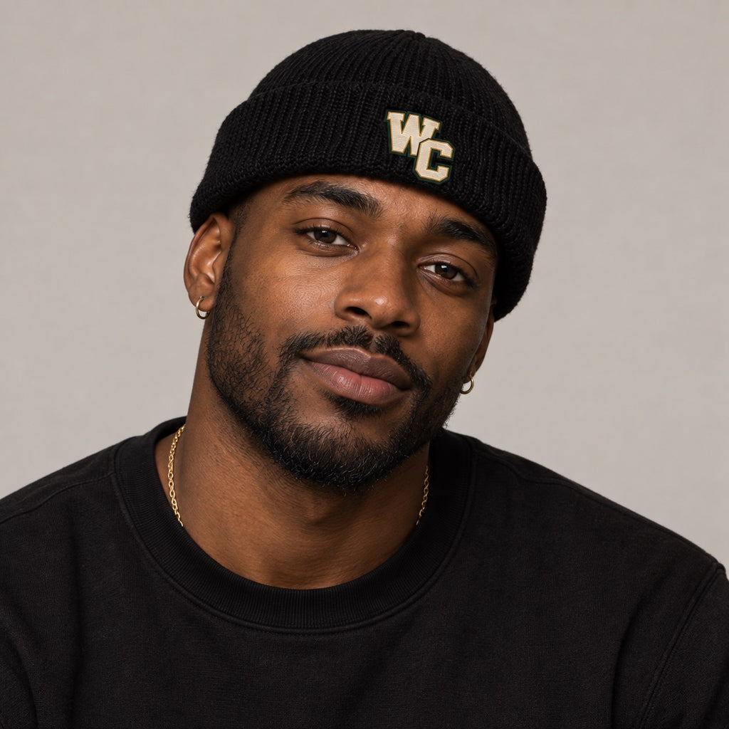 A man wearing a black knit beanie with gold embroidered letters 'WC' on the front, paired with a black crewneck sweatshirt and a gold chain necklace, photographed against a neutral gray background.